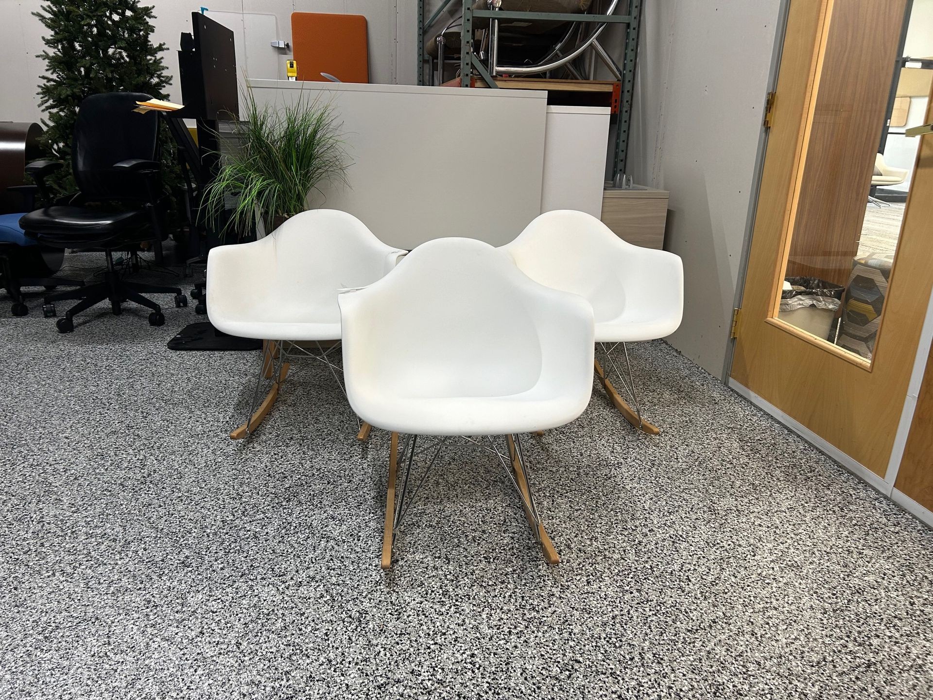 Three white rocking chairs on a speckled floor in an office with plants and miscellaneous furniture.